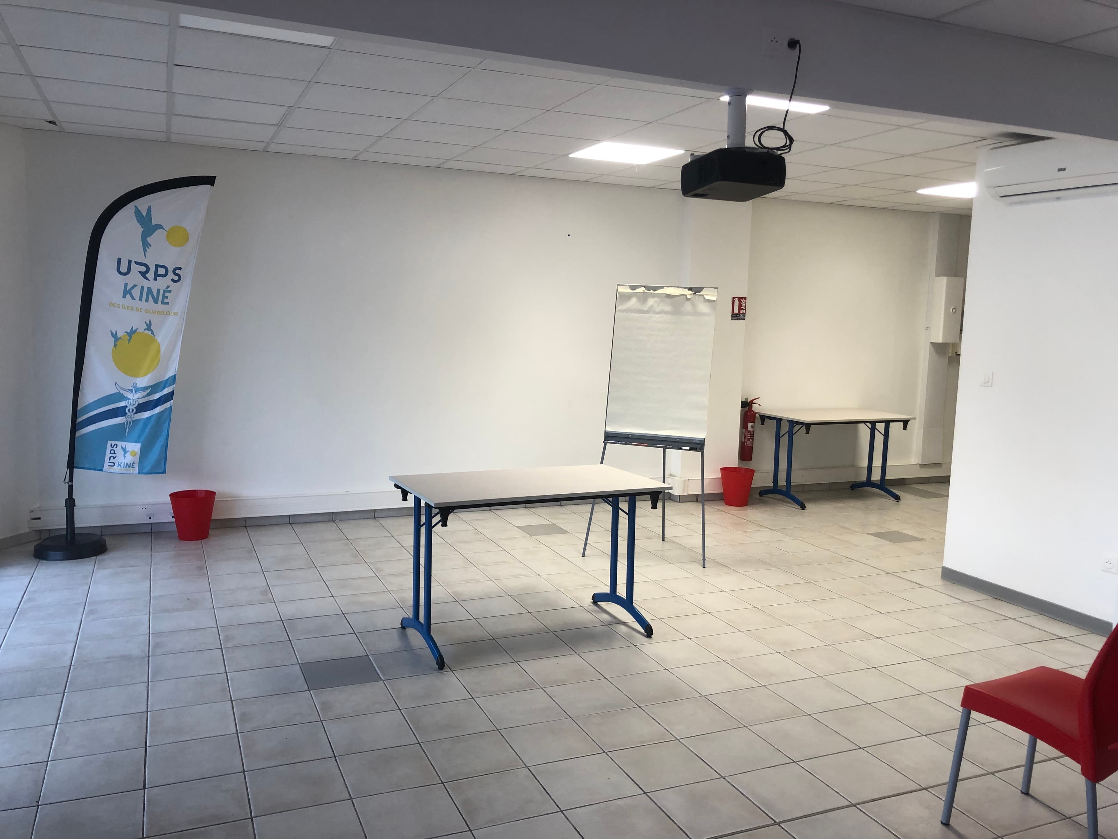 La salle vide, avec tables pliantes, paperboard et vidéoprojecteur au plafond